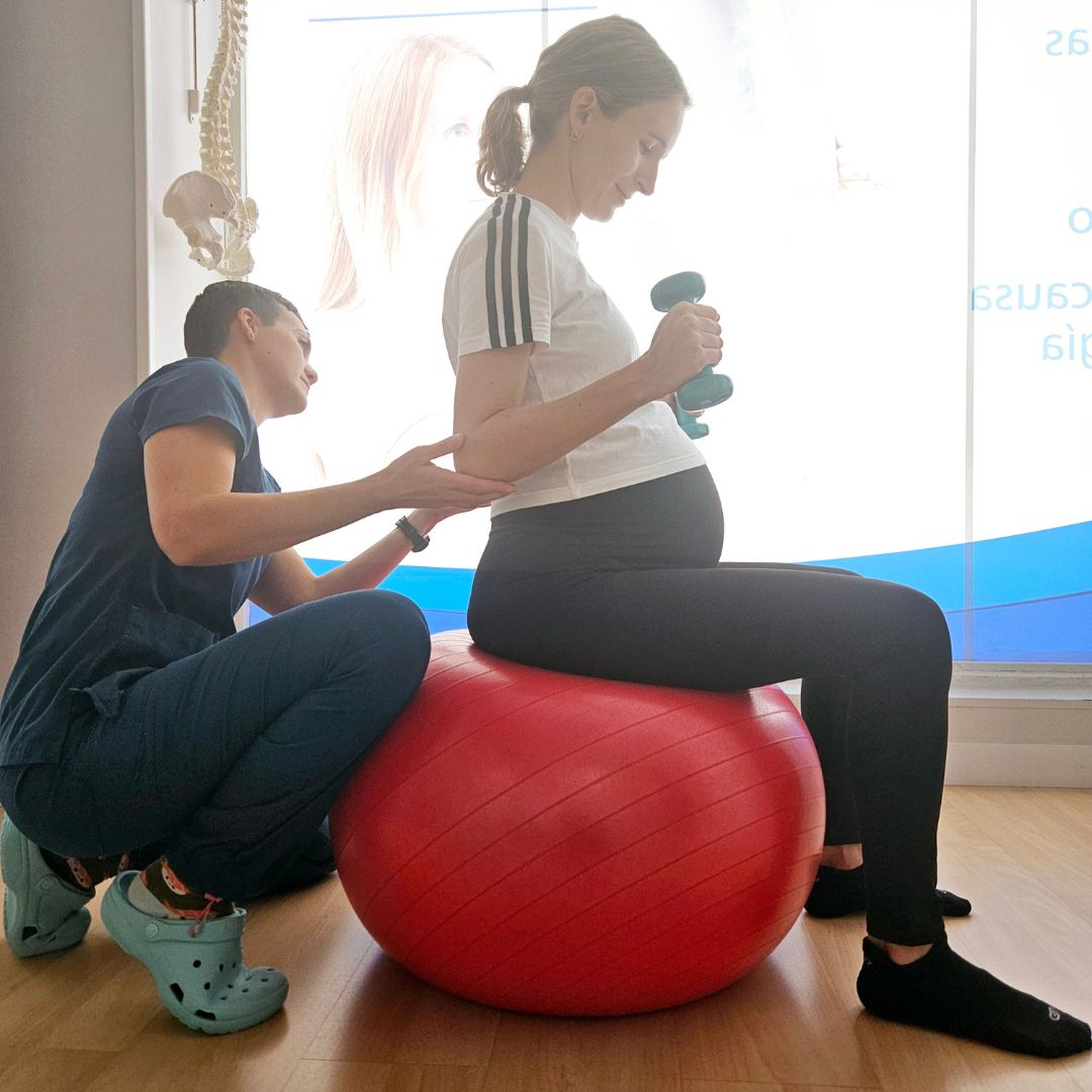 embarazada haciendo pilates sobre una pelota y con pesas mientras le corrige la fisioterapeuta